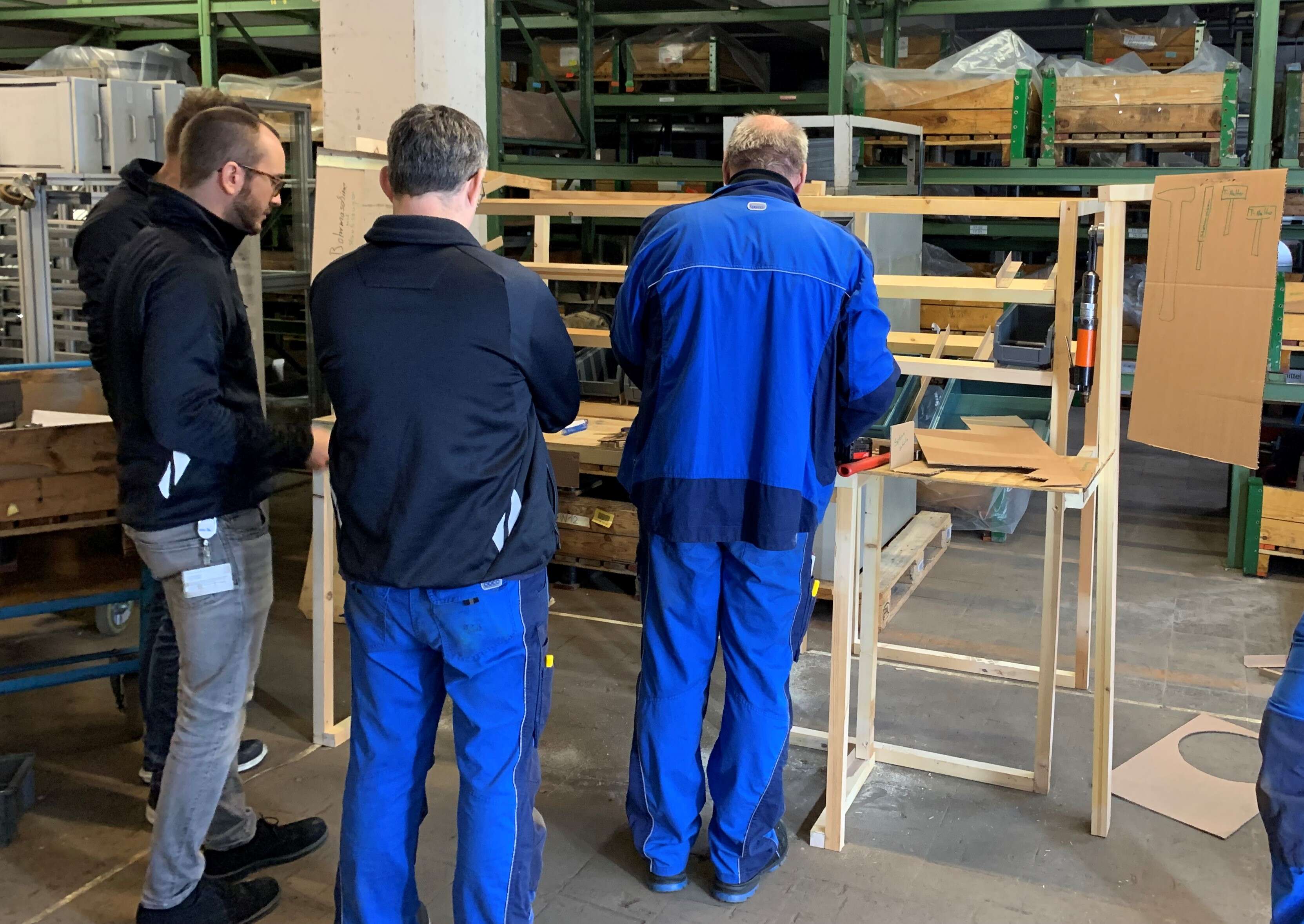 Drei Männer in Arbeitskleidung arbeiten in einer Werkhalle am Holzmodell eines Arbeitsplatzes.