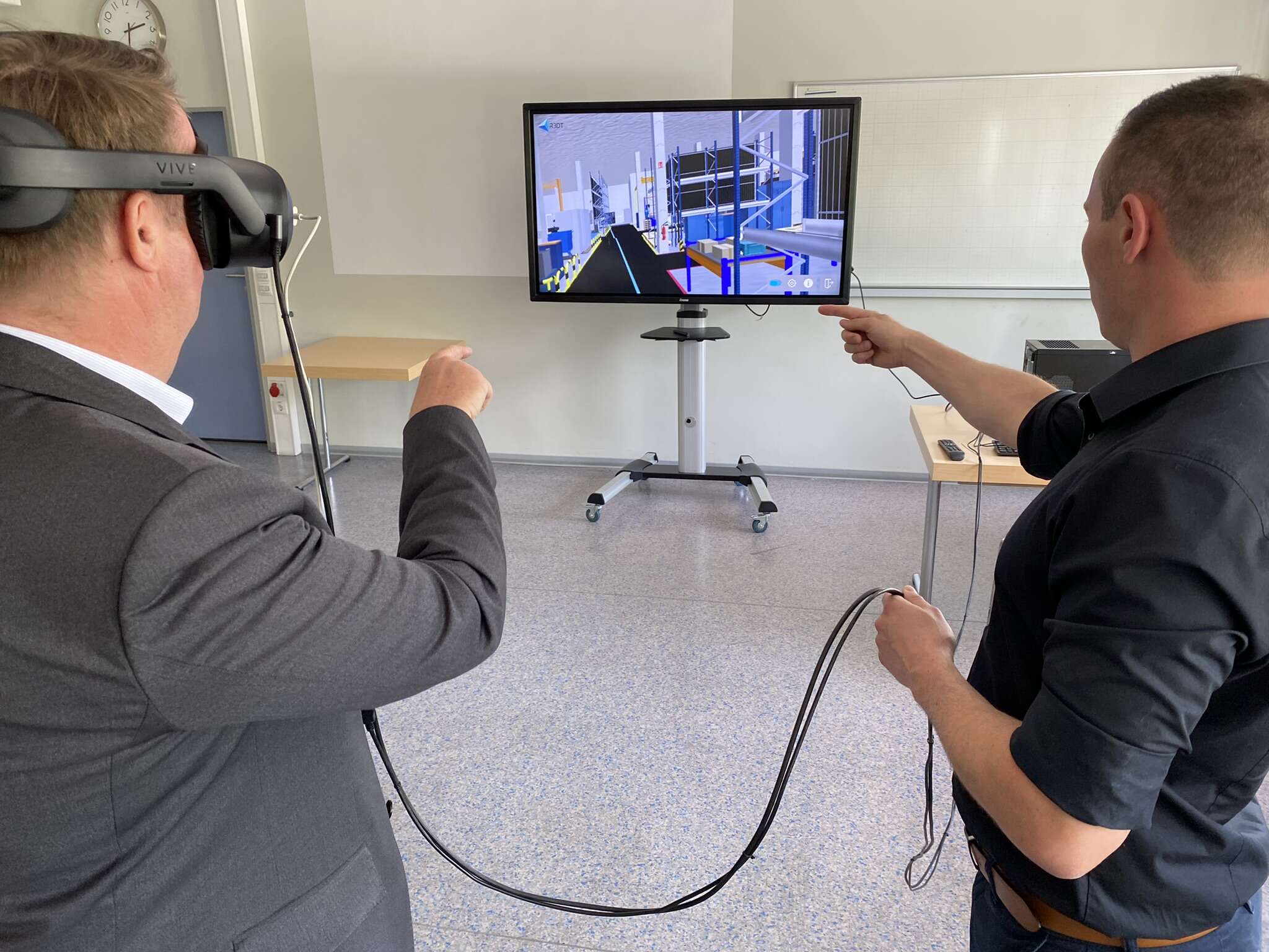 Two men, one of whom is wearing 3D glasses, are standing in front of a screen showing the virtual model of a production hall.