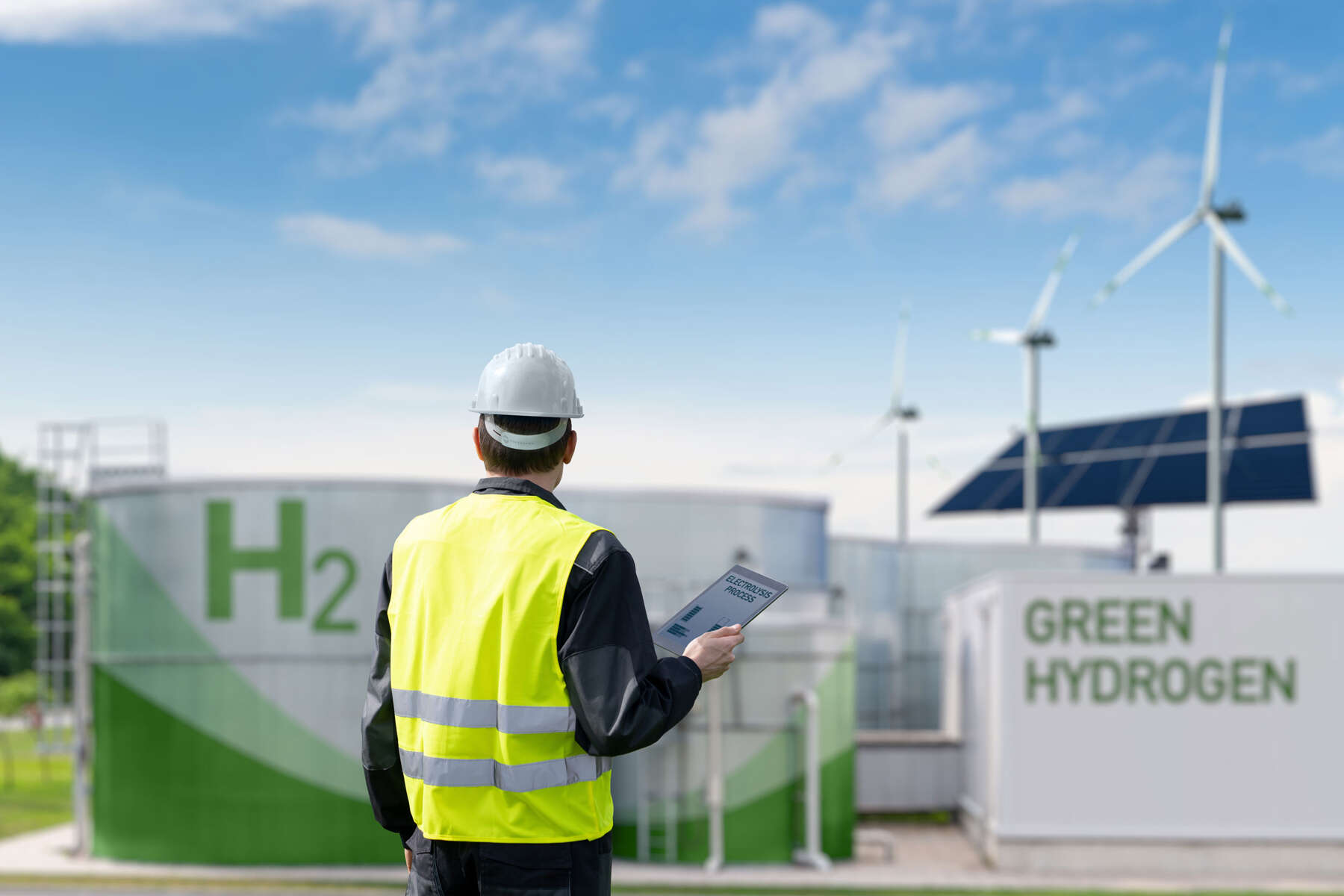 A man, wearing a hard hat and a yellow hi-vis vest, looking at hydrogen tanks, wind turbines and solar panels