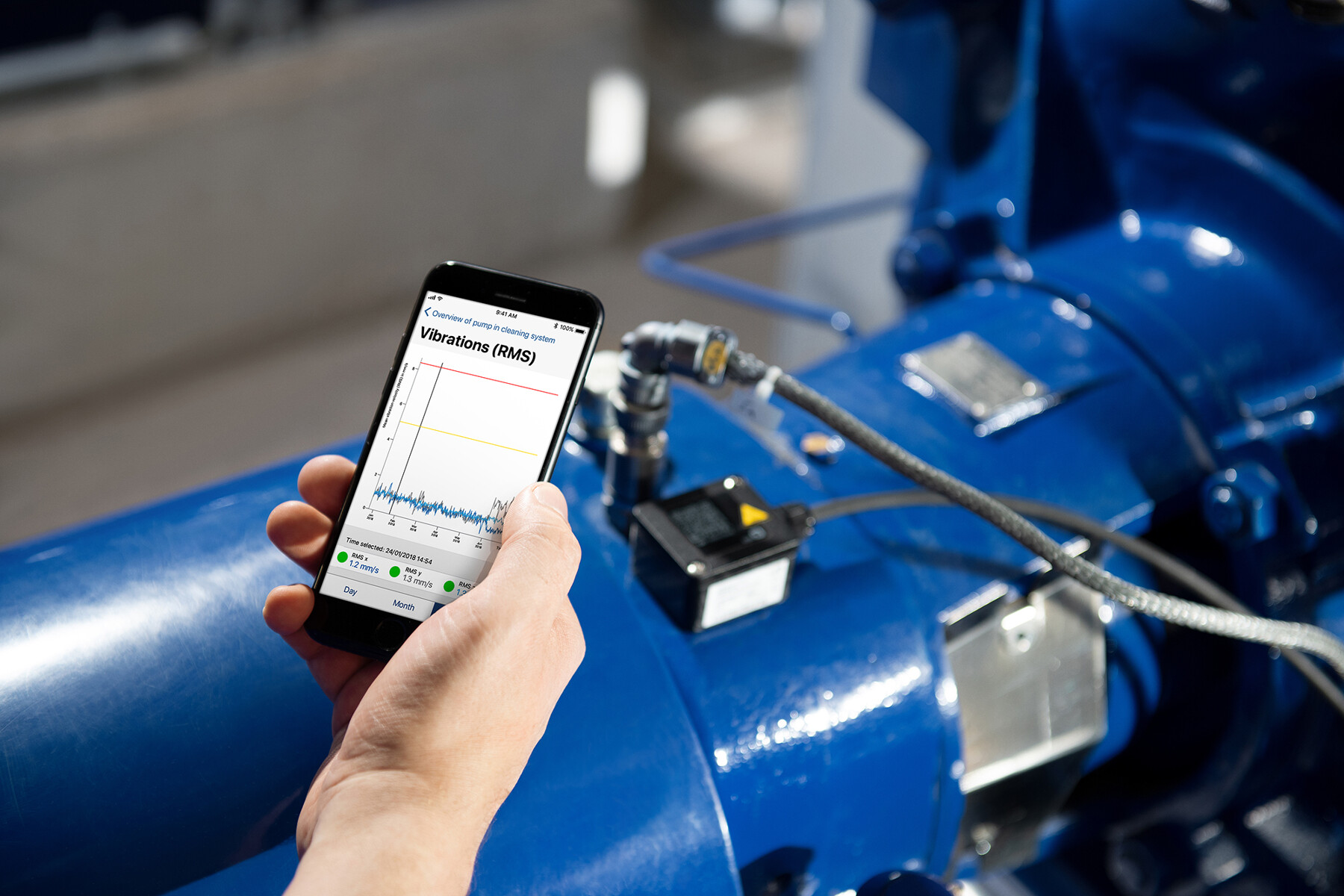 Hand holding a mobile phone with a graph displayed on the screen. In the background is a KSB pump with installed KSB Guard.