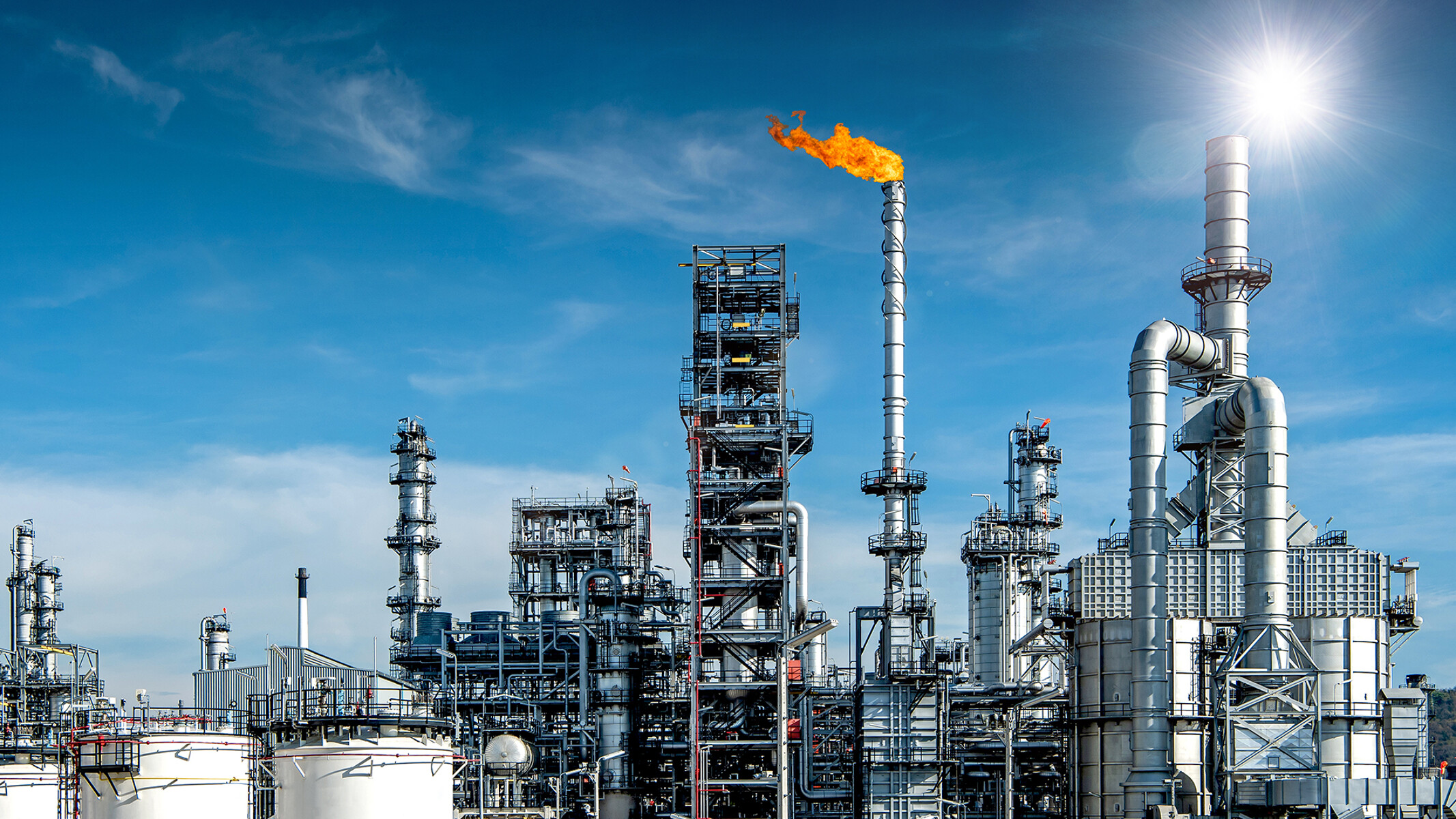 Velký komplex ropné rafinerie s oranžově zbarveným plynovým hořákem na pozadí zářivě modrého nebe