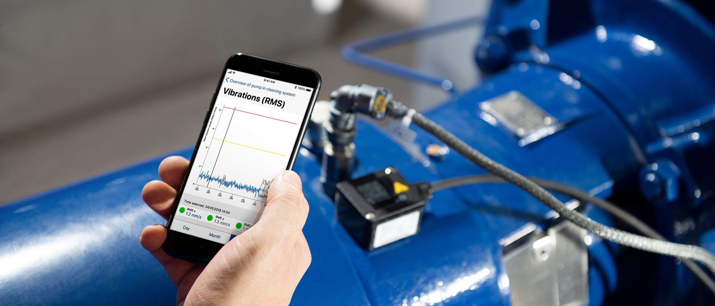 Hand holding a mobile phone with a graph displayed on the screen. In the background is a KSB pump with installed KSB Guard.