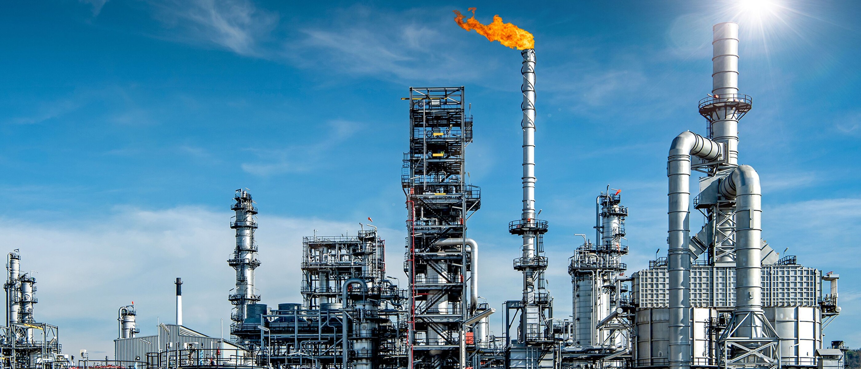 Gran complejo de refinado de petróleo con una llama de combustión naranja en el centro que contrasta con un brillante cielo azul